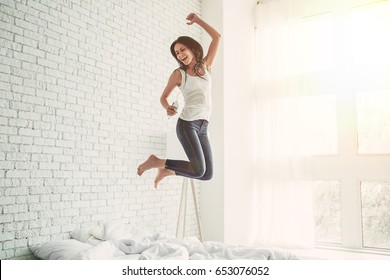 Happy young woman in earphones is listening to music with smart phone, jumping on bed, singing and smiling