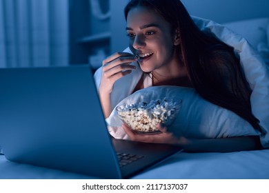 Happy young woman eating popcorn while lying on bed under blanket and watching movie on netbook at night at home - Shutterstock ID 2117137037