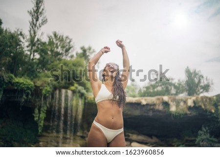 Happy young woman enjoying with arms up. She is in a tropical environment with waterfall in background. She is brunette. She is wearing sunglasses and white bikini