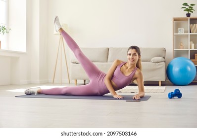 Happy Young Woman Enjoying Her Regular, Routine Fitness Workout At Home. Pretty Caucasian Girl In Pink Top And Yoga Pants Doing Side Lying Lateral Leg Raise On Rubber Exercise Mat In Living Room