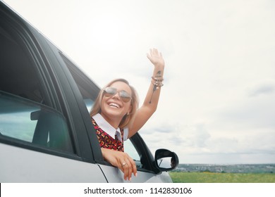 Happy  Young Woman Enjoying The Views On A Road Trip While Looking Through The Car Window, Saying Hello Waving Her Hand.