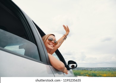 Happy  Young Woman Enjoying The Views On A Road Trip While Looking Through The Car Window, Saying Hello Waving Her Hand.