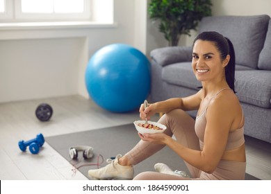 Happy Young Woman Enjoys Regular Fitness Exercise And Well-balanced Diet. Positive Smiling Female Sitting On Sports Mat After Routine Workout At Home, Eating Healthy Natural Food And Looking At Camera