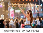 Happy young woman enjoys summer music fest, raises hands with peace sign, dances at beach party. Vibrant festival atmosphere, dusk light with stage lights. Fashionable crowd, holiday vibes, fun.