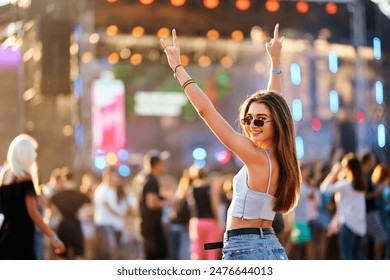 Happy young woman enjoys summer music fest, raises hands with peace sign, dances at beach party. Vibrant festival atmosphere, dusk light with stage lights. Fashionable crowd, holiday vibes, fun. - Powered by Shutterstock