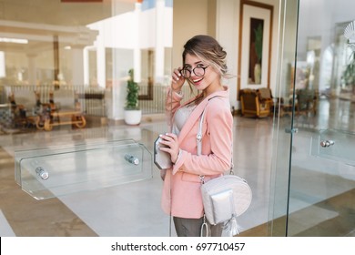 Happy Young Woman Entering Glass Door Into Modern Hotel, Cafe, Business Centre. Wearing Stylish Glasses, Pink Jacket, Little Silver Backpack.