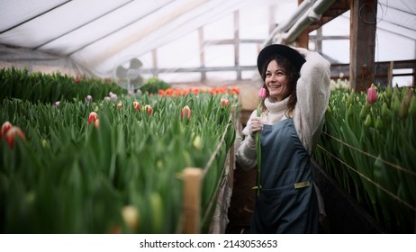 Happy young woman farmer in a hat sings song with tulip flower while working in a greenhouse