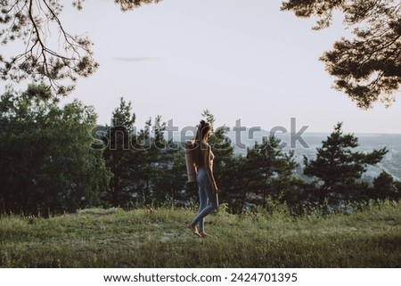 Happy young woman in fitness clothes with yoga mat going to workout in the forest at sunset.