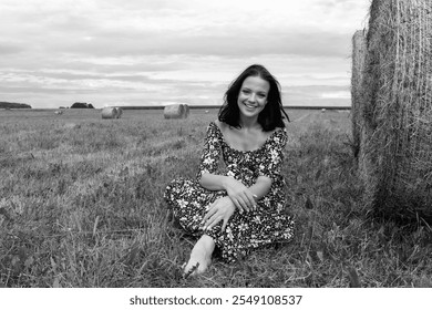 A happy young woman in a floral dress sits in a field with hay bales under a cloudy sky. Image contains noise and motion blur. Selective focus. Black and white image - Powered by Shutterstock