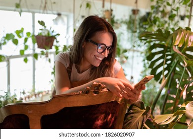 Happy young woman gardener in eyeglasses sitting on vintage chair in green house, holding smartphone, enjoying using mobile apps, chatting in social media, watching funny videos. Home gardening - Powered by Shutterstock