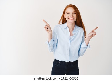 Happy Young Woman With Ginger Hair Pointing Fingers Sideways, Wearing Office Clothes And Standing Over White Background.