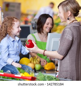 Happy Young Woman Giving Capsicum To Daughter With Cashier Smiling In Background At Super Market