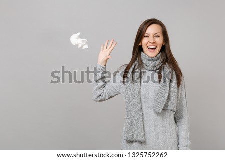 Happy young woman in gray sweater, scarf recovering and throwing paper napkin isolated on grey wall background. Healthy lifestyle, ill sick disease treatment, cold season concept. Mock up copy space