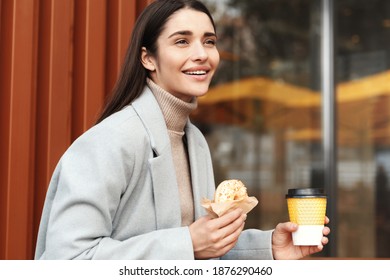 Happy Young Woman In Grey Coat Eating Donut In Coffee Shop, Drinking From Takeaway Cup And Smiling, Standing Outside Cafe.