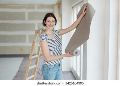 Happy young woman hanging wall paper while doing DIY home renovations standing in front of a newly painted section of wall holding a roll up on display - Powered by Shutterstock