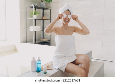 Happy Young Woman Has Fun Applying Face Toner With Cotton Discs. Gorgeous European Girl Wrapped In Towel After Bathing. Lady Is Sitting On Bathtub And Relaxing. Woman Takes Shower At Home.