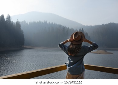 Happy Young Woman In Hat Enjoys Lake View In Mountains . Relaxing Moments In Forest. Back View Of Stylish Girl Enjoys The Freshness Outdoor. Freedom, People, Lifestyle, Travel And Vacation