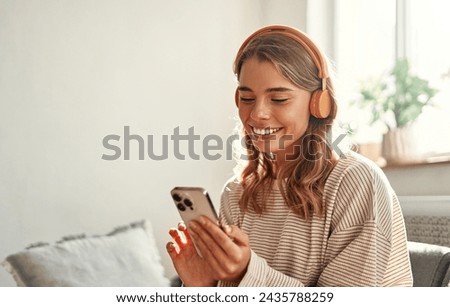 Happy young woman in headphones with phone on sofa in cozy living room at home relaxing and unwinding on weekend.