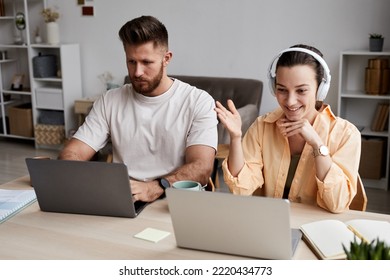 Happy Young Woman In Headphones Talking In Video Chat In Front Of Laptop While Sitting By Desk Next To Serious Bearded Man