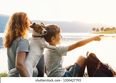 Happy Young Woman And Her Son With A Dog Jack Russell Terrier Are Sitting On The Bank Of The Lake In The Grass And Are Watching The Bright Sunset. Family Tourist Backpack. Weekend With Family. Camping