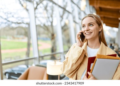 Happy young woman holding cellphone making call, smiling pretty blonde girl student talking on cell phone standing in university campus or coworking office advertising mobile plan. Copy space - Powered by Shutterstock