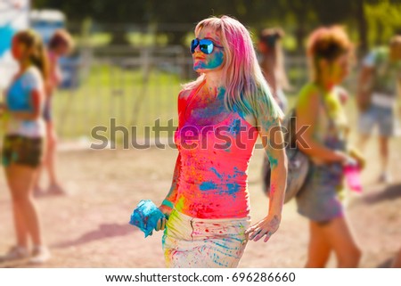 happy young woman holding colorful paint in hands and smiling with closed eyes while standing in field at holi festival