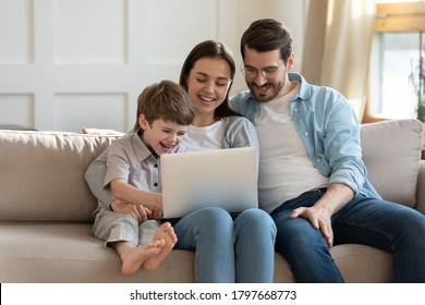 Happy Young Woman Holding Computer On Laps, Watching Funny Comedian Movie Video Or Shopping Online With Loving Husband And Adorable Little Child Son, Relaxing Together On Couch In Living Room.