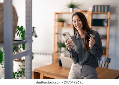 Happy young woman holding credit card using cell phone mobile app paying online making purchase in e-commerce digital store on smartphone at home. Virtual shopping, secure mobile banking concept. - Powered by Shutterstock