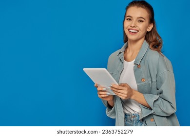 happy young woman holding digital tablet and smiling at camera on blue background, social media - Shutterstock ID 2363752279