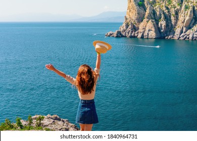 Happy Young Woman Holding A Hat And Running Down The Mountainside With A View Of The Rocks And The Sea. The Concept Of Home Away From Home