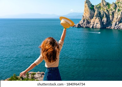 Happy Young Woman Holding A Hat And Running Down The Mountainside With A View Of The Rocks And The Sea. The Concept Of Home Away From Home