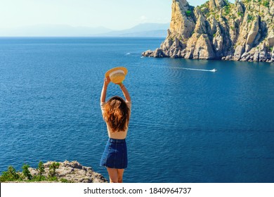 Happy Young Woman Holding A Hat And Running Down The Mountainside With A View Of The Rocks And The Sea. The Concept Of Home Away From Home