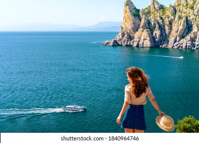 Happy Young Woman Holding A Hat And Running Down The Mountainside With A View Of The Rocks And The Sea. The Concept Of Home Away From Home