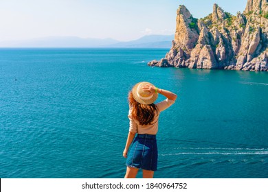 Happy Young Woman Holding A Hat And Running Down The Mountainside With A View Of The Rocks And The Sea. The Concept Of Home Away From Home