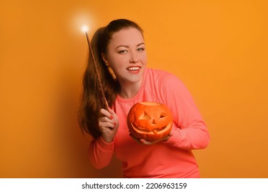 Happy young woman holding luminous magic wand or glowing magic stick with carved Halloween pumpkin or Jack o lantern pumpkin and looking at camera on yellow background.

Smiling girl is conjuring.