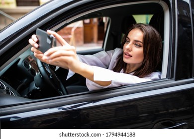 Happy Young Woman Holding Mobile Phone And Taking Photos While Driving A Car. Smiling Girl Taking Selfie Picture With Smart Phone Camera Outdoors In Car.