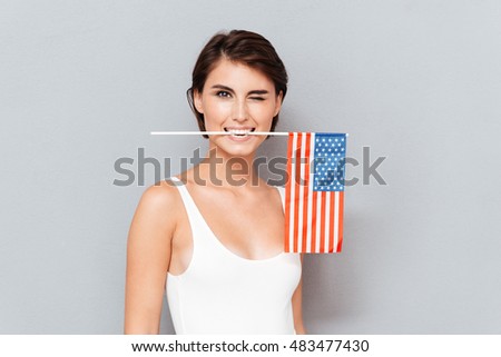 Happy young woman holding USA flag in teeth and winking over gray background