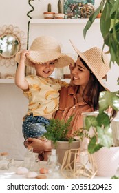 A Happy Young Woman Holds Her Daughter In Her Arms, They Laugh And Try On Straw Hats.Bright Cozy Home Interior With Indoor Plants.Family, Vacation, Summer Concept.