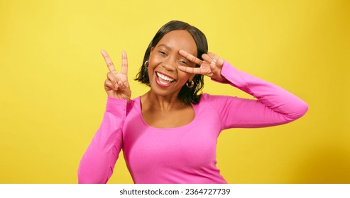 Happy young woman holds peace sign to eye, silly pose, yellow studio portrait - Shutterstock ID 2364727739