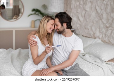 Happy young woman hugging her husband, holding positive pregnancy test on bed at home. Affectionate Caucasian couple planning their family, feeling excited to become future parents indoors - Powered by Shutterstock