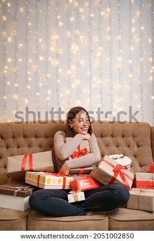 Happy young woman hugging many present boxes sitting crossed legs on a camel sofa with with lights in background