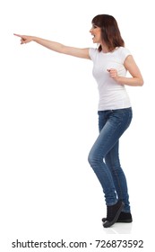 Happy Young Woman In Jeans, White T-shirt And Sneakers Is Holding Arm Outstretched, Pointing And Shouting. Side View. Full Length Studio Shot Isolated On White.