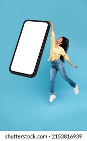 Happy young woman jumping up on blue studio wall, trying to catch reach and grab gadget, showing empty cell phone screen, full body length, collage. Excited lady recommending nice useful application - Shutterstock ID 2153816939