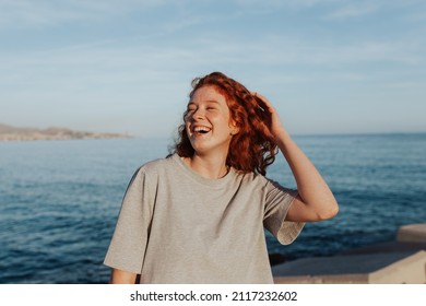 Happy Young Woman Laughing Cheerfully While Standing Next To The Ocean. Carefree Redhead Woman Having A Good Time In The Summer Sun. Young Woman Enjoying A Serene Weekend By The Seaside.