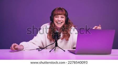 Happy young woman laughing while recording a live radio show in a studio. Cheerful young woman hosting an audio broadcast in neon purple light. Young woman creating content for her internet podcast.