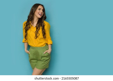 Happy young woman in linen shirt and shorts is posing with hands in pockets, looking to the side and smiling. Three quarter length shot against blue background. - Shutterstock ID 2165505087