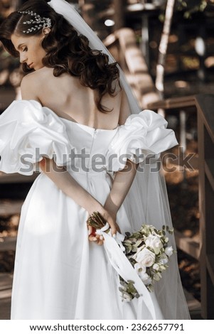Happy young woman with long curly hair in a white wedding dress, holding a bouquet of flowers, outdoors. Beautiful girl with flowers. Vertical photo