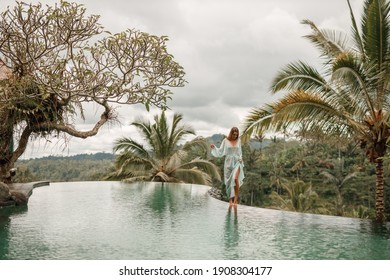 Happy young woman in long  dress walking on the edge of swimming pool and enjoying summer vacation in daytime