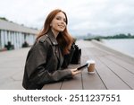 Happy young woman with long red hair and leather jacket enjoying coffee break standing on riverfront promenade, taking moment holding mobile phone in hands. Female smiling in calm outdoor setting