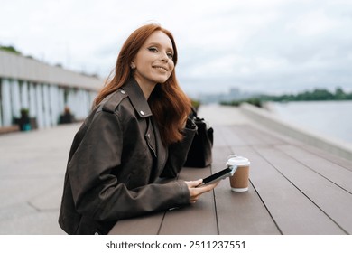 Happy young woman with long red hair and leather jacket enjoying coffee break standing on riverfront promenade, taking moment holding mobile phone in hands. Female smiling in calm outdoor setting - Powered by Shutterstock